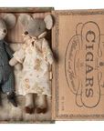 Two Maileg Mice by Maileg, one in blue checkered pajamas and one in a floral dress, rest side by side in a vintage-style cigar box with a printed label—a delightful scene from the Maileg Mouse collection.