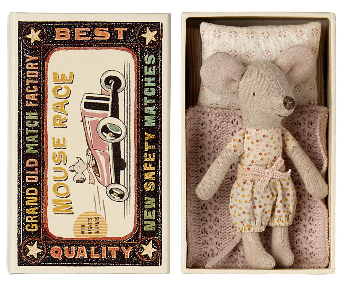 A Maileg Mouse in polka-dot pajamas rests in a small bed with a knitted blanket, tucked inside a matchbox-style box. The Maileg-branded box lid displays vintage text and an image of a mouse driving a red car.