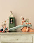Five Maileg toy mice, including the Maileg Tricycle Mouse, Big Sister, ride bikes and tricycles down a cushion ramp on a plush bear, with magnets securing the scene atop a vintage-style dresser near a dollhouse cabinet and lamp.