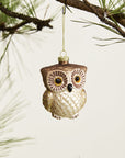 The Accent Decor Owl Ornament, crafted in brown and cream glass, hangs from a gold string on a pine branch, creating a charming forest accent against a simple background.