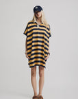 A person stands against a plain background wearing the Denimist Rugby Shirt Dress in yellow and navy stripes, crafted from cotton, with hands in the dress pockets. The look is styled with brown boat shoes and a navy cap with white lettering.