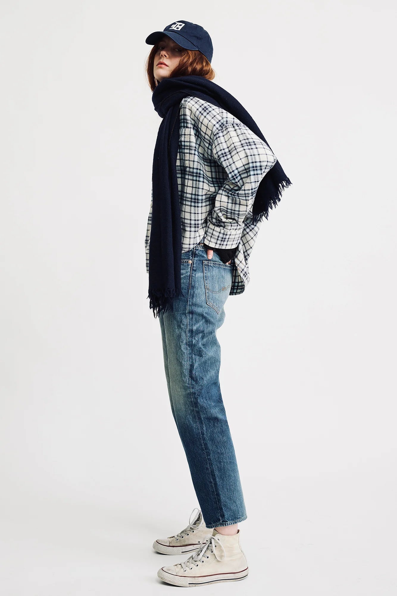 A person in a plaid shirt, Denimist LUCY BF JEAN ESHER BLUE with a relaxed fit, white socks, and white sneakers stands sideways. They wear a navy scarf and cap, with brown hair showing, against a plain white background.