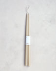 The Floral Society 18" Taper Candle - Pair in beige features a classic design with a single looped wick for hanging. Expertly crafted, they come bundled with a white paper label and rest on a light grey textured surface.