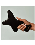 A hand holds The Body Gua Sha—a uniquely shaped black massage tool by Faire, crafted from bian stone and engraved with "wild method." The serene, neutral background evokes balance.