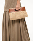 A person in a beige dress holds the Staud Lena Raffia Clutch, featuring brown leather handles and a rectangular trim.