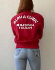 A person with long hair wears a 1980s Alpha Cubic Racing Jacket by Red Vintage Co., featuring "ALPHA CUBIC RACING TEAM" on the back and a loose boxy fit, paired with light blue ripped jeans against a plain light-colored wall.