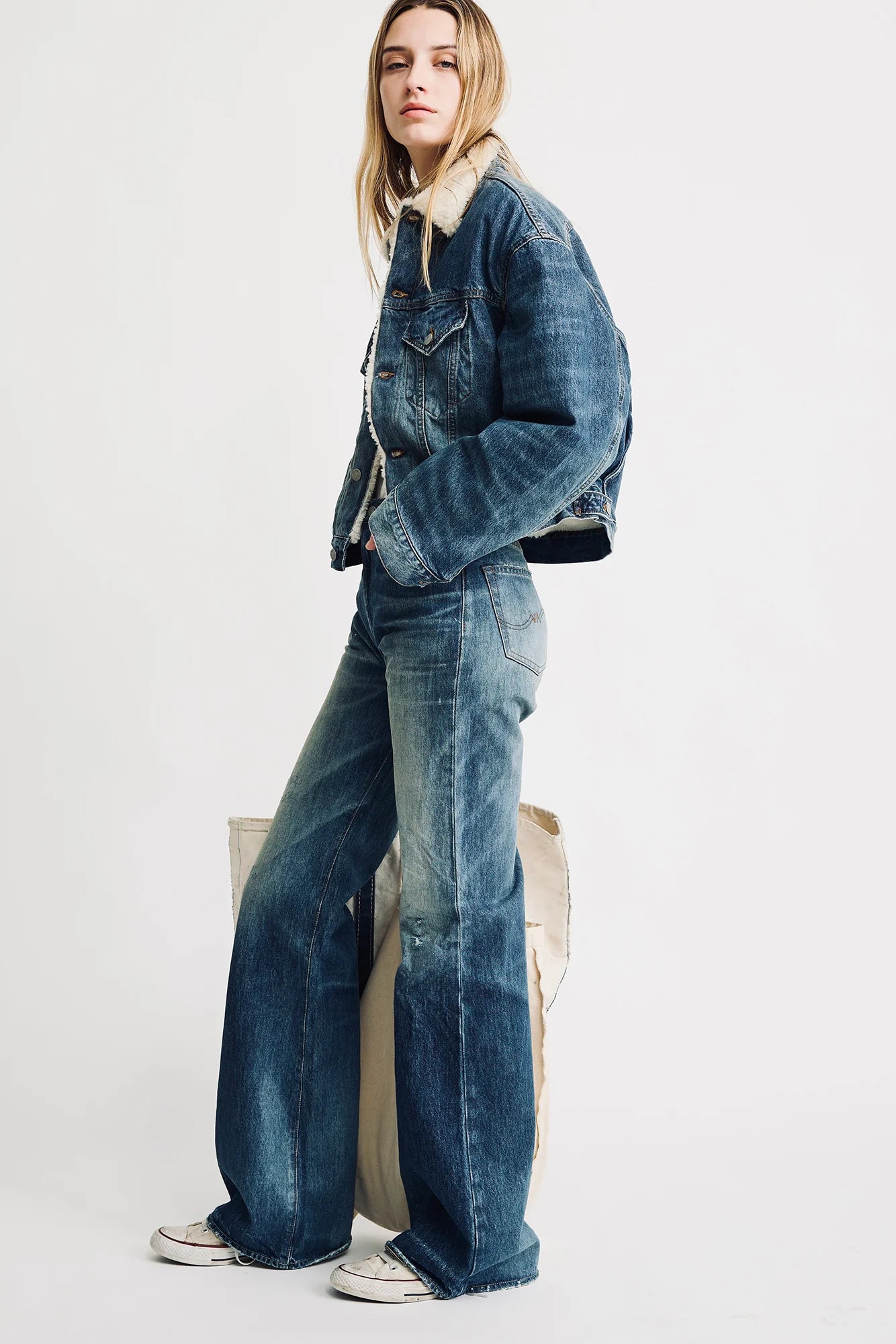 A person poses with hands in pockets, wearing the Denimist IRIS BOXY TRUCKER JACKET against a plain background, styled with flared blue jeans and white sneakers, and displaying a neutral expression.
