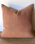 The Fringe Accent Pillow Havane 22x22, featuring a square shape and textured fabric in muted dusty rose, sits upright on a wooden surface against a plain white wall.