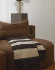 A cozy, brown curved sofa features the elegant Jitana Wholesale Thiago Momo Blanket, with its black and beige stripes offering charm. Behind, a minimalist white vase with dark foliage rests on a brown pedestal. The room's neutral tones and soft lighting echo refined sustainable practices.