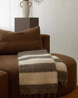 A cozy brown sofa showcases the Thiago Momo Blanket by Jitana Wholesale, expertly crafted by Mayan artisans with dark brown, light brown, and cream stripes. In the background, a tall vase with dried branches sits on a pedestal in a well-lit and inviting setting.