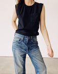A person in Hey Gang's The Muscle Tee and blue jeans stands against a white background, one hand in their pocket and a small forearm tattoo visible.