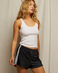 A woman with long blonde hair wears the black Mikayla Travel Short by Perfectwhitetee, featuring white side stripes, and a lightweight white tank top while standing before a beige fabric backdrop.