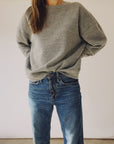 A person is wearing "The Boxy Sweatshirt" from Hey Gang, featuring an oversized vintage fit in a loose gray style, paired with high-waisted blue jeans that have visible buttons. The sweatshirt is slightly tucked into the jeans, and the indoor setting with a plain background emphasizes the unisex appeal of this cozy ensemble.