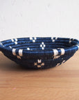 The Kabaya Small bowl by Amsha - Faire, a shallow round sisal woven basket with blue and white stripes, sits on a light wooden surface against a white textured wall.