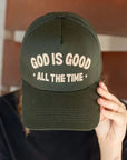 A person with braided hair, partially visible from the front, is wearing a dark green "God is Good Hat" by Faire with the embroidered text "God is good all the time." The person's hand is holding the brim of the hat, slightly covering their face, against a blurred background.