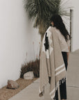 A woman with long dark hair walks beside a white wall and desert plants, draping the Faire TT Handwoven Blanket Taos Two over her shoulder.