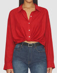 A person wears Denimist's SLIM TWISTED FRONT BEACH SHIRT in red, styled with blue high-waisted jeans and a black belt. The cropped image highlights the shirt’s twisted front and frames the shoulders and hips.