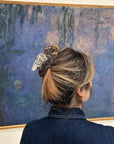 A person with light brown hair in a bun, secured with Faire's Hand Painted Pigeon Clip, stands before an impressionist painting of water lilies in soft blue-purple hues.