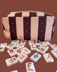 The Faire Wild West Mahjong SET—including tiles, 4 racks, a bag, and mat—is displayed on a brown surface with scattered Western Mahjong tiles showing unique symbols and illustrations.