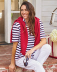 A smiling woman with long brown hair sits on brick steps, wearing the Frank & Eileen Patty Popover SS Henley, white pants, a red sweater over her shoulders, and holding sunglasses. A cream tote bag and white flowers sit beside her.