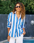 A woman with wavy hair wears sunglasses and the Frank & Eileen Eileen 26 blue-and-white striped relaxed fit shirt with white pants, standing by a pool in front of a gray wall and lush greenery.