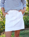 A person stands outdoors with hands in the utility pockets of a white Frank & Eileen Jameson Skirt, paired with a long-sleeve navy and white striped shirt. Greenery and flowers create a softly blurred background.