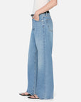 A person wearing FRAME’s The Pixie Loose light blue wide-leg jeans with a petite inseam, a white sleeveless top, and black open-toe sandals stands with one hand in their pocket against a plain white background.