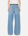A person is seen from behind wearing FRAME's The Pixie Loose jeans—light blue, rigid denim with visible back pockets and a relaxed wide-leg fit. Only the lower back and legs are visible; the jeans are paired with a white top.