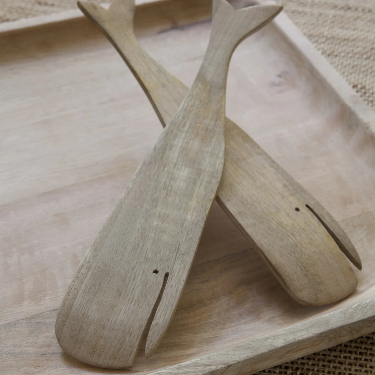 The Sydney Whale Serving Set by Be Home features two handcrafted, whale-shaped salad servers with carved details, made from sustainably sourced mango wood, crossed on a light tray atop a textured surface.