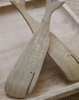 The Sydney Whale Serving Set by Be Home features two handcrafted, whale-shaped salad servers with carved details, made from sustainably sourced mango wood, crossed on a light tray atop a textured surface.