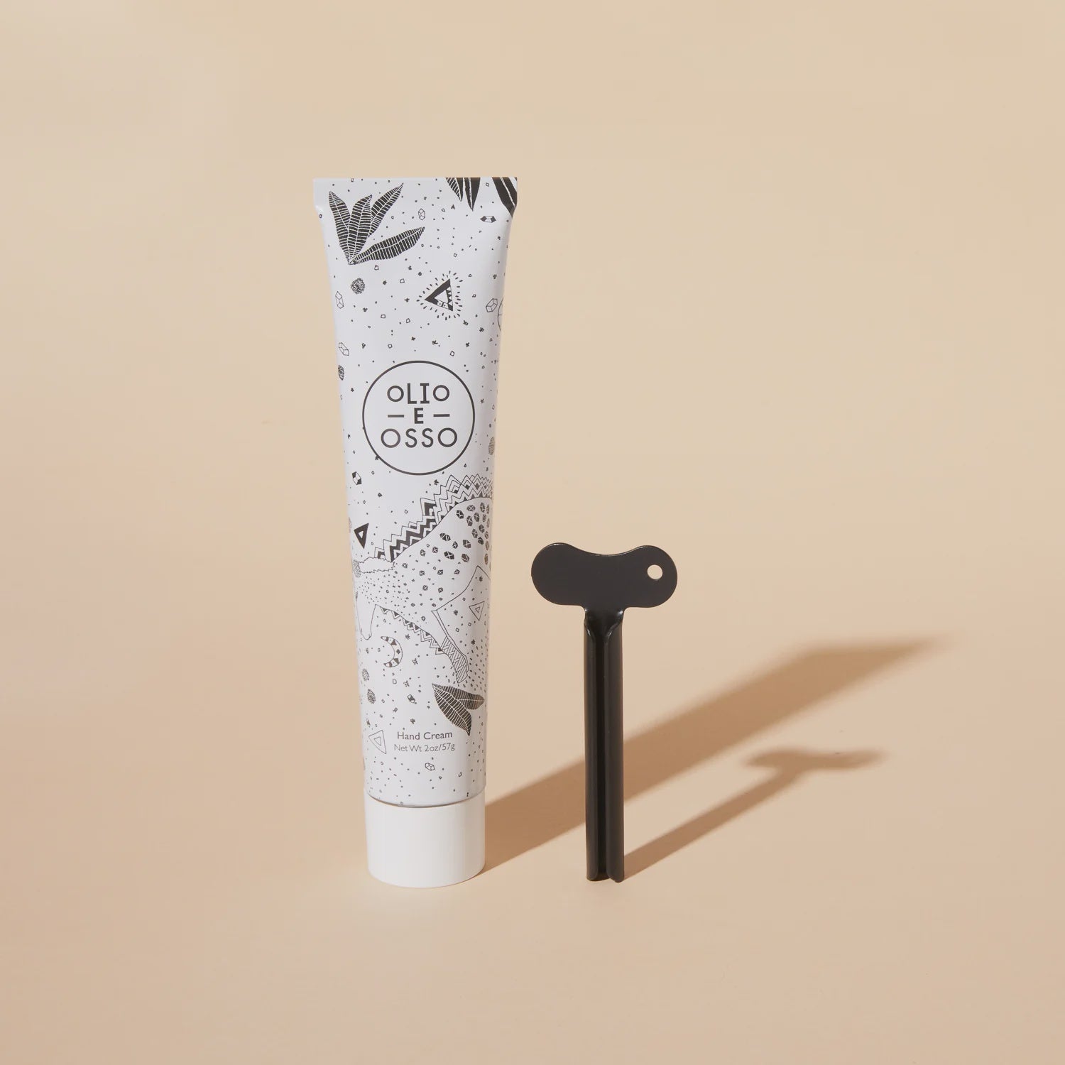 A tube of Olio E Osso hand cream placed next to a black T-shaped applicator on a light beige background. The tube features a stylish black and white geometric pattern inspired by Scottsdale Arizona.