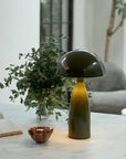 The Leoma Lamp Olive by Abigail Ahern sits on a marble surface beside a vase of greenery, a small decorative bowl, and an open book. In the background, there’s a gray chair and a large window.