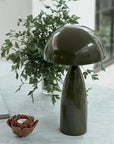 The Leoma Lamp Olive by Abigail Ahern, with its glossy dark green dome shade, is displayed on a marble table alongside a brown ceramic candle holder, an open book, and a glass vase filled with leafy branches in a bright room.