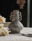 A small, speckled Abigail Ahern Marilla Vase with decorative handles sits on a textured surface beside cream flowers and scissors, set against a soft, blurred background.