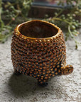 The Abigail Ahern C13-004 Tortoise Small Pot, a textured ceramic planter with a bumpy brown finish and tortoise head detail, sits on stone with greenery blurred in the background.