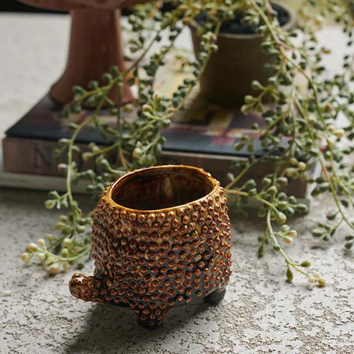 The Abigail Ahern C13-004 Tortoise Small Pot, a textured brown ceramic planter with three legs and a tail-inspired handle, rests on stone, framed by leafy greenery and a stack of books in the background.