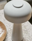 The Abigail Ahern Adelaide LED Lamp White, a modern minimalist table lamp with a round flat shade and conical base, sits on a textured surface near a wooden plate. It's dimmable, portable, and USB rechargeable for convenience.