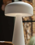 The Adelaide LED Lamp White by Abigail Ahern, a modern mushroom-shaped portable table lamp, glows softly in a warmly lit room with blurred natural elements and a window in the background.