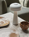 The Adelaide LED Lamp White by Abigail Ahern sits on a textured stone table near two rustic wooden plates and a brown ceramic cup, with a green upholstered chair in the background.