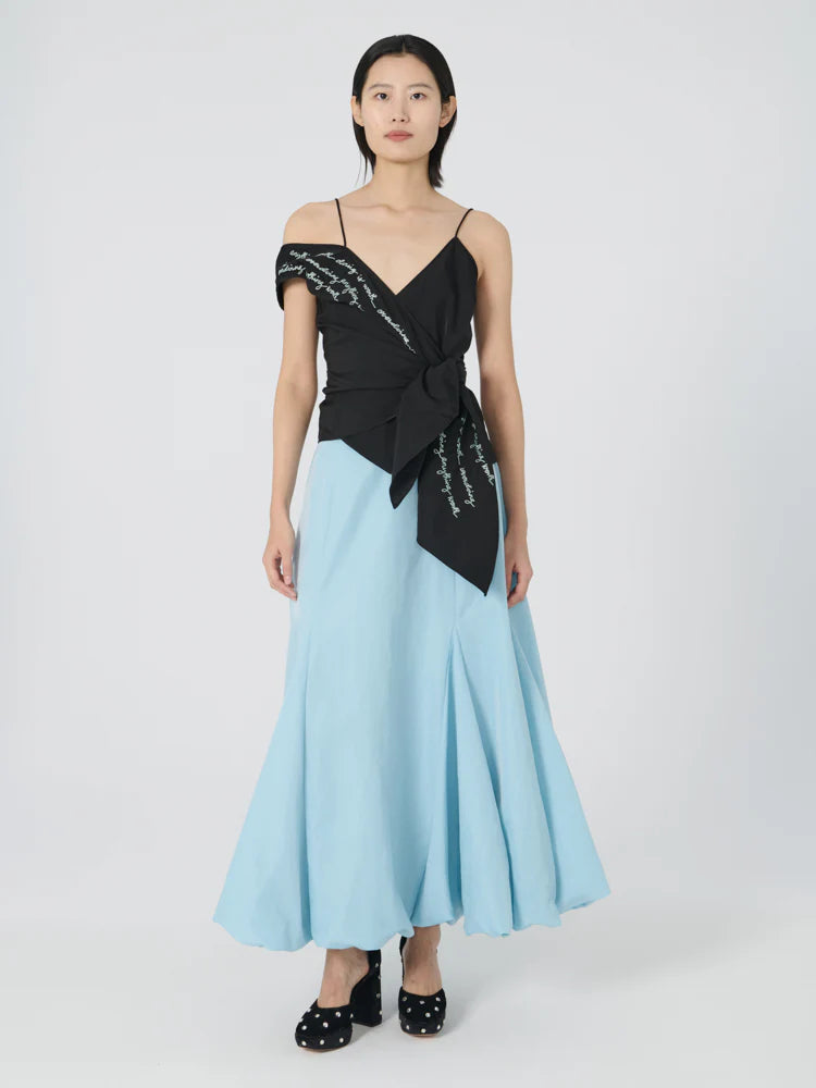 A person stands against a plain background wearing a stylish ensemble: an asymmetric black top featuring embroidered wording and a bow, paired with the Donna Bubble Skirt from Lingua Franca in a light blue hue. The outfit is completed with black polka dot shoes. The person's hair is straight and shoulder-length.