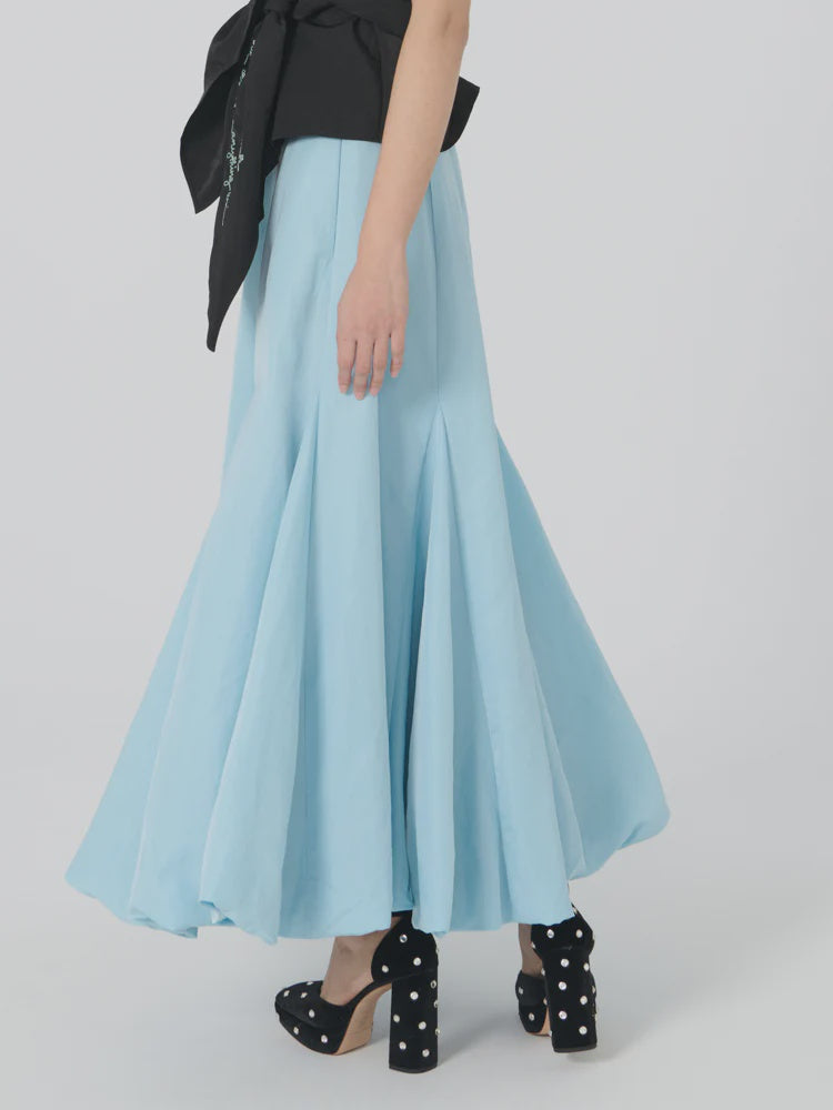 A person wearing the Donna Bubble Skirt by Lingua Franca in a flowing light blue hue is captured mid-step. They pair it with a black sleeveless top featuring a large bow, and walk confidently in black high heels embellished with white studs against a plain white backdrop.