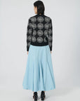 A person with shoulder-length dark hair is adorned in a black sweater showcasing an embroidered phrase, paired elegantly with the Lingua Franca's Donna Bubble Skirt in a long, flowing light blue hue. They face away from the camera against a simple white background, effortlessly combining style and understated elegance.