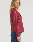 A person with long blonde hair, wearing JAKETT's Allison Burnished Leather red blazer and blue jeans, stands sideways against a plain light background.