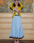 A woman stands smiling with her hands on her head, wearing a yellow sweater adorned with abstract patterns, the Donna Bubble Skirt by Lingua Franca in a light blue hue that flows gracefully, and pink shoes. The backdrop features a painted floral design on a wooden wall.