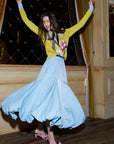 A woman joyfully spins in the Donna Bubble Skirt by Lingua Franca, a long and flowing light blue skirt featuring a velvet waistband. She pairs it with a bright yellow sweater adorned with colorful patterns, standing in front of a decorative background highlighted by gold curtains. Her hair cascades loosely around her shoulders, and she completes the look with pink shoes.
