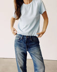 A person dressed in Hey Gang's The Crew T-shirt in enzyme-washed light blue and 100% cotton jeans adjusts the waistband, standing against a plain backdrop with their head out of frame.
