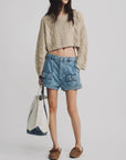 A woman wears the Denimist Cropped Cable Sweater with denim shorts, a blue bucket hat, brown loafers, and a large canvas bag—modern proportions update this classic knit against a plain light background.