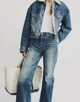 A woman wears the Denimist IRIS BOXY TRUCKER JACKET in indigo denim with sherpa lining, paired with matching high-waisted flared jeans and a white tee. She carries a large off-white tote; her hair is loose and her face partly out of frame.