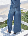 Wearing Frank & Eileen’s Madeline Patch Pocket Pant and sandals, a person stands on a white wooden deck overlooking the beach and greenery, capturing a relaxed coastal vibe.