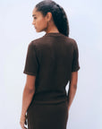 A woman with braided hair faces away, wearing the Luxe Cotton T-shirt by White + Warren in dark brown with a matching skirt. She accessorizes with gold hoop earrings and a bracelet against a light blue background.